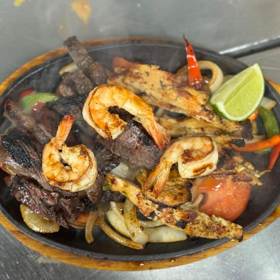 Sizzling mixed grill platter with shrimp, grilled steak, chicken, vegetables, lime wedge, and red pepper on a cast-iron serving plate.