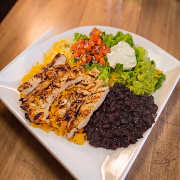 Grilled chicken strips served with yellow rice, black beans, lettuce, guacamole, pico de gallo, and sour cream on a white plate.